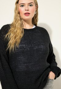Schwarzer Strickpullover mit einem strukturierten Muster, Rundhalsausschnitt und lockerer Passform, getragen über einem weißen Oberteil, was zu grauen Jeans passt.