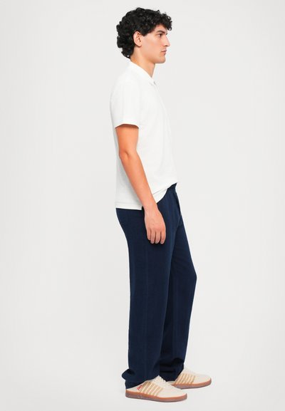 Weißes Polohemd mit kurzen Ärmeln, marineblaue, locker sitzende Hose und helle Sneakers mit gestreiften Akzenten, von der Seite vor einem hellen Hintergrund gesehen.