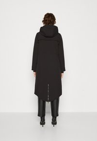 Manteau long noir avec capuche, ourlet incurvé et fermeture éclair à l'arrière. Caractérisé par une texture lisse et un design minimaliste. Associé à un pantalon noir.