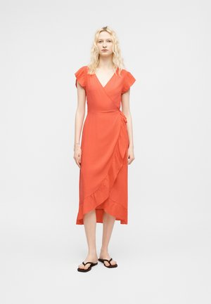 Tânără cu păr blond ondulat, purtând o rochie coral înfășurată cu tiv cu volane și șlapi negri, stând pe un fundal simplu, deschis la culoare.