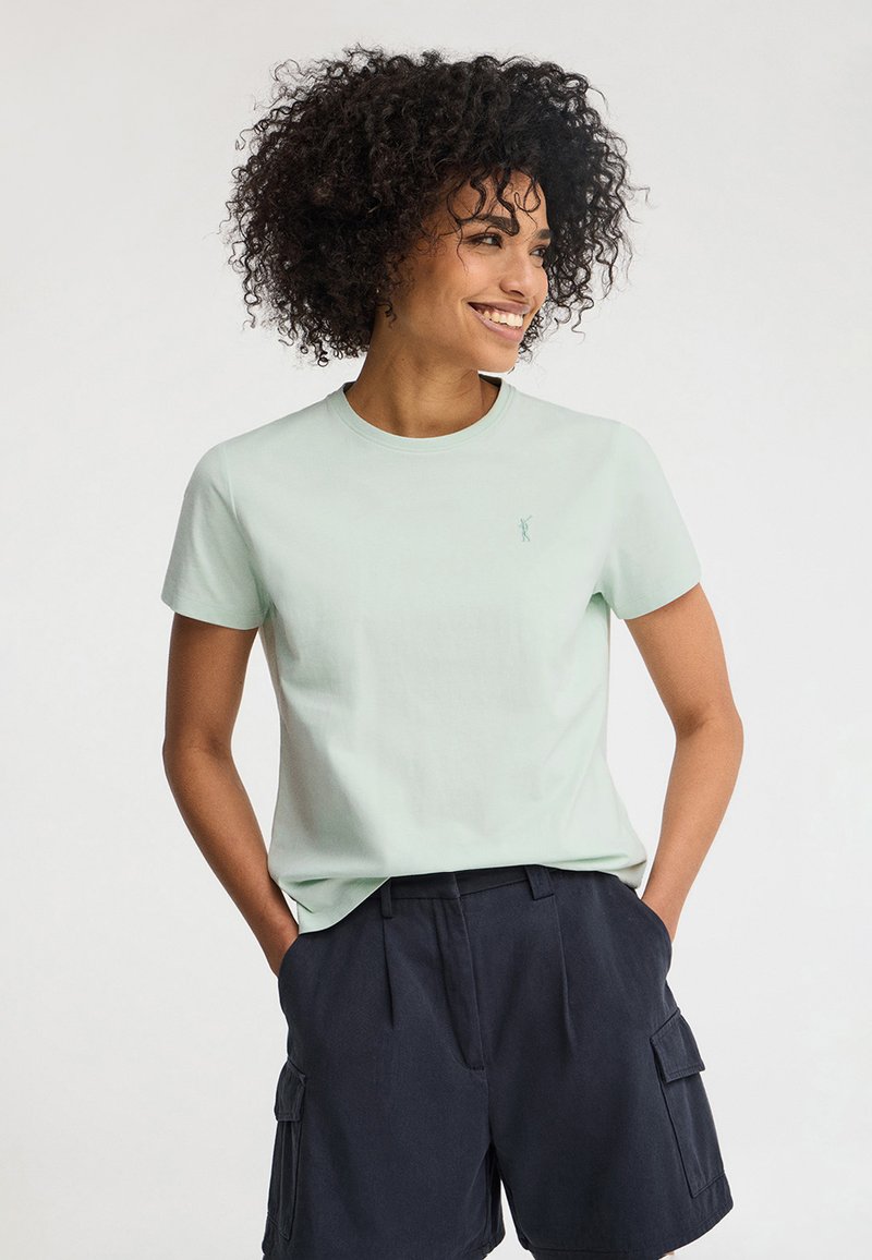 Mujer sonriente, vistiendo una camiseta de manga corta verde claro y pantalones cortos cargo azul marino oscuro, de pie con las manos en los bolsillos frente a un fondo liso.