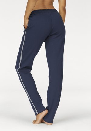 Bareback person standing on one foot wearing navy blue pants with light grey side stripes and elastic waistband, hands in pockets.