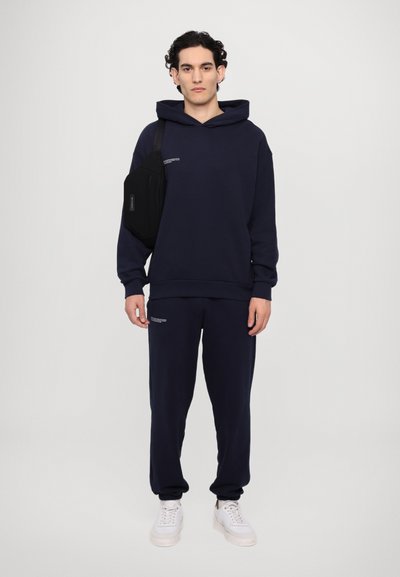 Helleblauer Kapuzenpullover und passende Jogginghose aus weichem Stoff, mit einer Kängurutasche und Textdetails, kombiniert mit weißen Sneakern und einer schwarzen Umhängetasche.