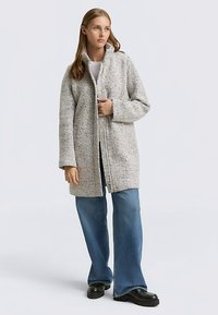 Grau strukturierter Reißverschluss-Mantel mit hohem Kragen, lockerem Sitz und großzügigen Taschen, getragen über einem weißen Oberteil und blauen Schlagjeans.