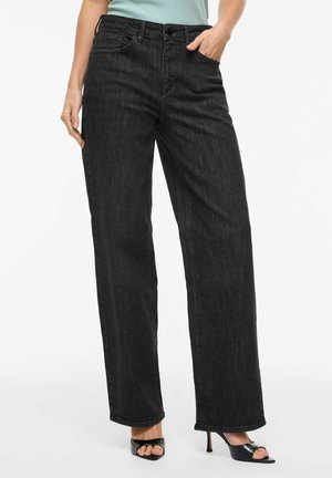 Ženska v črnih kavbojkah z visokim pasom in ravnimi nogavicami, kombiniranimi z odprtimi črnimi sandali s peto in svetlo modro prilegajočo majico.