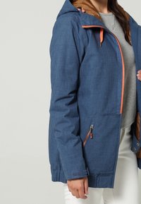 Niebieska kurtka z teksturą, z pomarańczowym zamkiem błyskawicznym i akcentami. Posiada kaptur i boczne kieszenie, wyściełane brązowym materiałem. Tkanina wydaje się lekka.