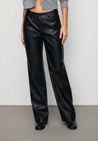 Kvinde iført høj-taljede sorte bukser i kunstlæder med krokodillemønster og sorte sko med firkantet tå, cropped grå sweater synlig over taljen.