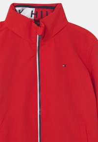 Rode jas met een hoge kraag, ritssluiting aan de voorkant en zijzakken. Voorzien van een klein logo in marineblauw en wit op de linkerborst. Soepele textuur.