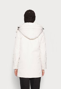 Biała izolowana kurtka z kapturem, o gładkiej fakturze i wydłużonym kroju. Widoczne zamki błyskawiczne i detale przeszyć na rękawach.