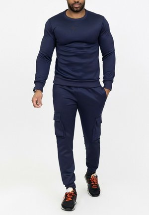Homme marchant, portant un sweat-shirt à manches longues bleu marine et un pantalon de jogging cargo, avec des baskets noires ornées de lacets orange.