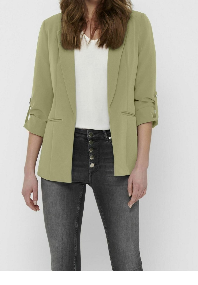 Femme portant un blazer vert clair avec manches retroussées, un haut blanc et un jean noir taille haute avec fermeture à boutons, debout devant un fond uni.