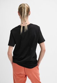 Personne avec trois tresses blondes porte une chemise noire à manches courtes et un pantalon corail, debout les mains dans les poches, dos à la caméra.