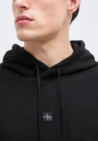 Noor mees, kes kannab musta Calvin Kleini kapuutsiga pusa lipsude ja kapuutsiga, kujutatud lõuast rindkereni ühevärvilise tausta ees.