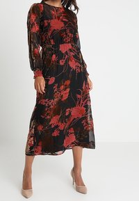 Lang sort kjole med rødt blomstermønster, gennemsigtigt stof, elastisk talje og lange ærmer. Kombineret med nude højhælede sko.