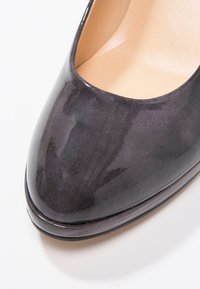 Chaussure de ville pour femme en cuir verni noir avec bout arrondi et doublure intérieure beige sur fond blanc.