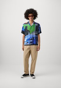 Short-sleeve, multicolored shirt featuring graphic prints of cars and a cat. Worn with beige trousers and black sneakers.