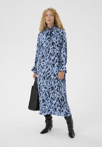 Femeie cu păr creț, purtând o rochie midi cu buline albastre și negre, cu guler înalt și cravată, cizme negre, ținând o geantă tote neagră, stând pe un fundal alb.