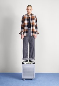 Model w brązowej, czarnej i białej koszuli w kratę flanelową, nosi ją na czarnej koszulce, w połączeniu z szarymi szerokimi spodniami i czarno-białymi trampkami, stojąc na srebrnym ruchomym pudełku.