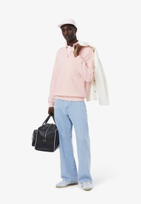 Maglione rosa chiaro con zip a un quarto, jeans a gamba larga blu chiaro, giubbotto di jeans bianco, borsa da viaggio nera, sneaker bianche e un cappellino rosa.