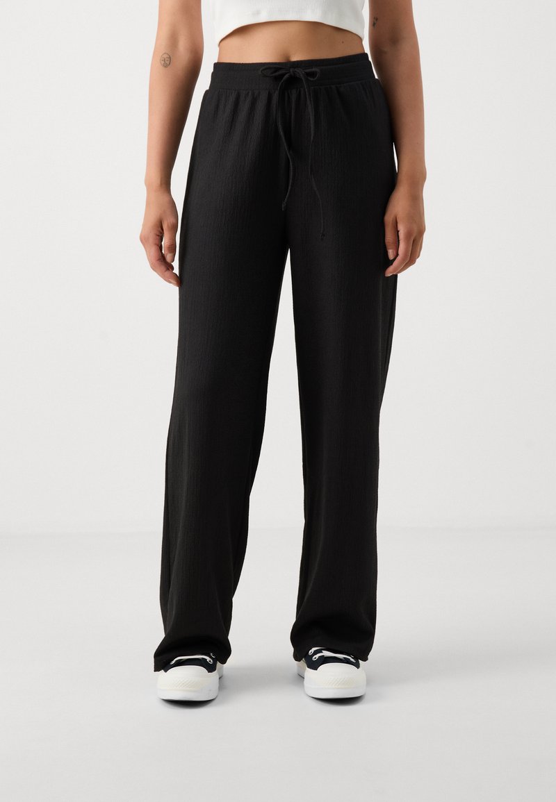 Zwarte wijde pantalons van textielstof, met een elastische tailleband en een trekkoord. Draagt met witte sneakers.