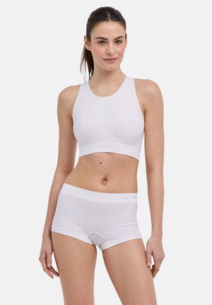 Jeune femme portant un soutien-gorge de sport blanc sans couture et un shorty assorti, debout devant un fond clair uni.