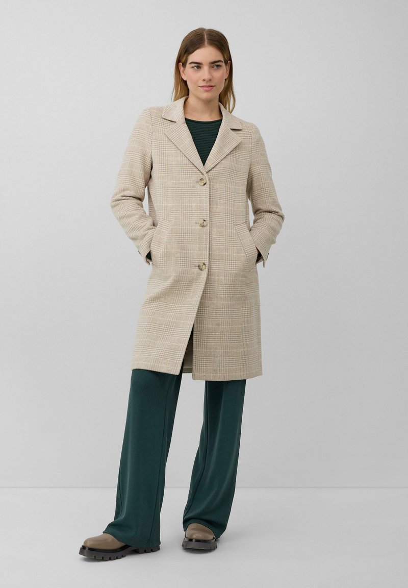 Vrouw in een beige geruite knie-lange jas, donkergroene wijde broek en beige schoenen met dikke zool, staand met handen in de zakken.