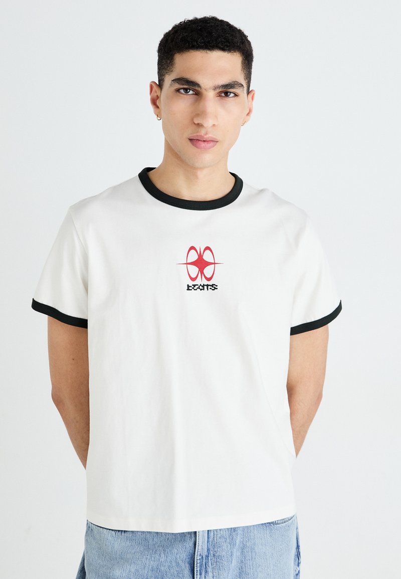 White cotton t-shirt with black ribbed neck and sleeve hems. Features a red graphic design centered on the chest with stylized text underneath.