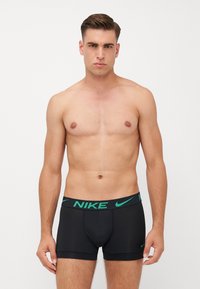 Črne bokserice z zelenim Nike logotipom na pasu, izdelane iz gladkega, elastičnega materiala, z oprijetim dizajnom in dolžino do sredine stegen.