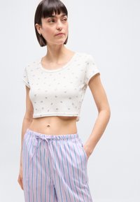 Femme aux cheveux courts et foncés portant un crop top blanc avec de petits motifs de papillons et un pantalon ample à rayures violettes, regardant sur le côté.