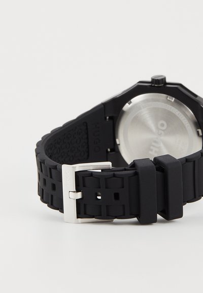Bracelet de montre en caoutchouc noir avec texture géométrique, boucle en acier inoxydable et fond de boîtier rond en acier inoxydable argenté. Logo de la marque visible.