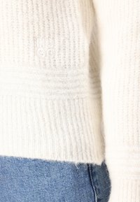 Biały sweter dziane z ryżowym splotem, z subtelnym haftowanym logo, połączony z niebieskimi dżinsami. Miły i przytulny wygląd.