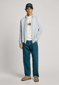 Light blue linen shirt, white graphic t-shirt, dark blue denim jeans, beige sandals, navy cap with turquoise text, standing against a neutral background.