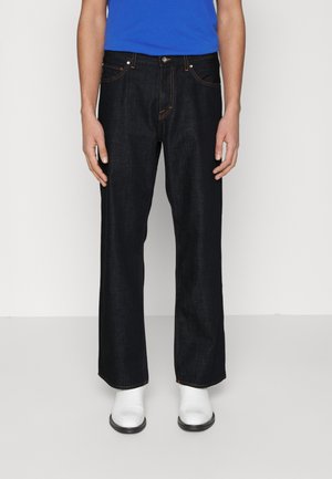 Osoba nosząca ciemne, proste dżinsy, niebieską koszulę i białe buty, stojąca na jednolitym jasnym tle.