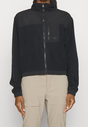 Personne portant une veste polaire noire zippée avec une poche poitrine et un pantalon beige, debout devant un fond gris uni.