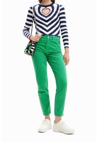 Kobieta w zielonych spodniach, białych sneakersach, pasiastym topie z długim rękawem z sercowym wycięciem, niosąca torbę na ramię w czarno-białym wzorze.