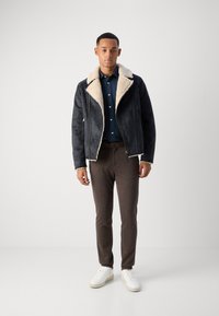 Black faux suede jacket with cream shearling collar, navy button-up shirt, brown patterned trousers, and white sneakers.