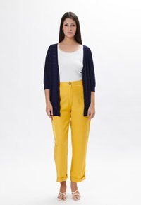 Cardigan blu navy, top bianco, pantaloni gialli con una texture liscia e risvolti, abbinati a sandali bianchi con cinturini.