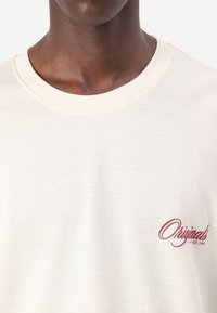 Kreemikas puuvillane T-särk, millel on ribatud ümardatud kaelus ja väike punane "Originals" logo vasakul rinnal. Pehme tekstuur.