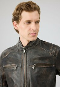 Bruine leren motorjack met een opstaande kraag, rits aan de voorkant en twee ritszakken op de borst. Gepresenteerd met een gestructureerde afwerking en subtiele kleurvariaties.