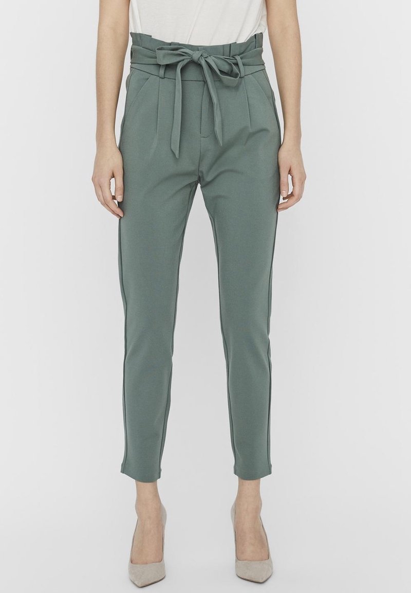 Femme portant un pantalon vert taille haute et fuselé avec une ceinture en tissu nouée, associé à des talons pointus beige clair et un haut blanc.