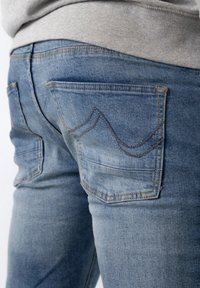 Nahaufnahme von blauen Denim-Jeans mit sichtbaren Gesäßtaschen und oranger Naht, getragen mit einem hellgrauen Oberteil.
