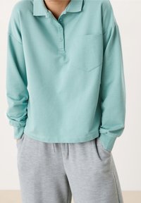 Camisa de polo de manga larga color verde menta con cuello, tres botones y un bolsillo en el pecho, combinada con pantalones deportivos texturizados de color gris claro.