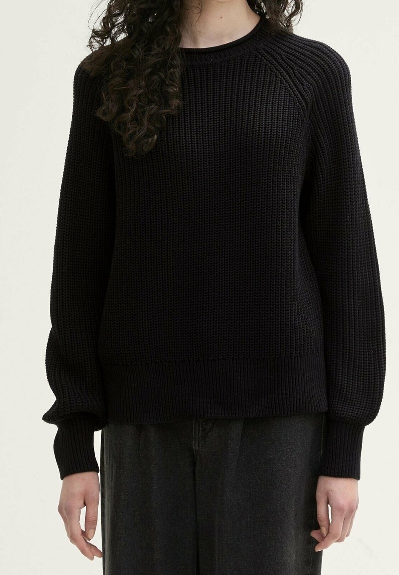 Pull noir en maille avec un motif côtelé texturé et un col rond. Il présente des manches longues et un ourlet ajusté. Porté avec un pantalon foncé.