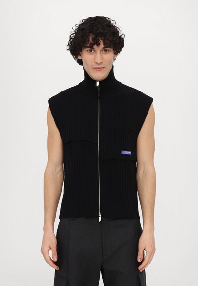 Top senza maniche nero a coste con colletto alto, chiusura frontale con zip e dettaglio del logo blu a contrasto sulla sinistra, indossato da un modello.