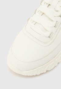 Λευκό αθλητικό παπούτσι με υφή στην επιφάνεια, στρογγυλεμένη μύτη και ραμμένες λεπτομέρ�ειες. Διαθέτει επίπεδα κορδόνια και παχιά ελαστική σόλα με λεπτομέρεια λογότυπου.