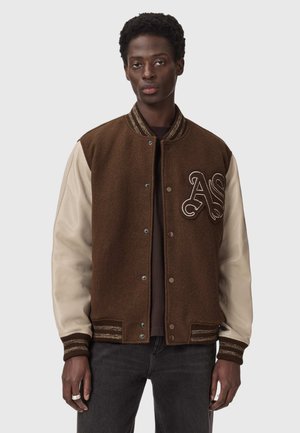 Mand iført brun varsityjakke med beige læderærmer og broderede "AS" initialer på brystet, kombineret med mørke jeans.