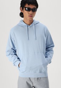 Felpa in cotone azzurro chiaro con tasca frontale, coulisse regolabile e polsini a coste. Il modello indossa occhiali da sole neri e pantaloni grigi.