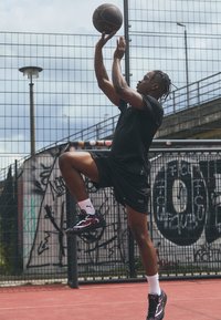 Un joueur de basket-ball noir, vêtu d'une tenue de sport noire, saute pour tirer un ballon de basket marron. Il porte des baskets noires et violettes ainsi que des chaussettes blanches.