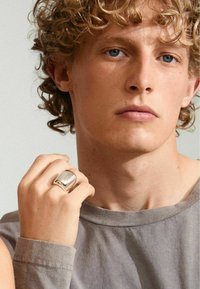 Anillo de plata pulida con un diseño suave y redondeado, llevado en una mano, junto a una camiseta gris de manga corta con cuello redondo.