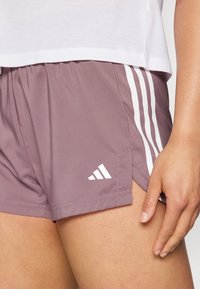 Detailní záběr na osobu nosící mauve sportovní šortky s bílými pruhy a malým logem Adidas, spolu s bílým tričkem.